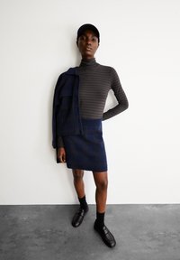 Marineblauer, plissierter Rock mit strukturiertem Muster, kombiniert mit einem grau gestreiften Rollkragenpullover und einer dunklen Jacke lässig über die Schulter gelegt. Schwarze Schuhe und eine Mütze.