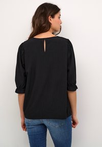 Blusa nera con righe verticali testurizzate, maniche a sbuffo a tre quarti e un piccolo dettaglio a occhiale sulla parte posteriore. Indossata con jeans blu.