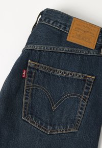 Poche arrière d'un jean en denim bleu foncé avec des coutures jaunes et une étiquette rouge Levi's, plus un patch en cuir beige Levi Strauss & Co. sur la ceinture.