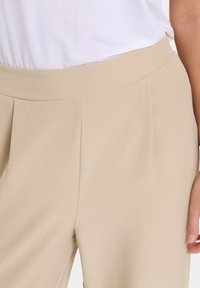 Gros plan sur un pantalon taille haute beige avec des plis, porté avec un haut blanc rentré, montrant une partie d'une main sur le côté.