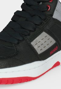Baskets noires avec lacets noirs, panneaux gris, accents rouges et semelle blanche avec relief rouge, montrant une partie du nom de la marque "HUGO."