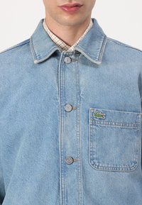 Light blue denim jacket with gray buttons, a single front pocket, and a small green crocodile logo. Collared with a checked shirt underneath.