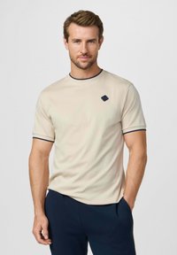 Man draagt een beige T-shirt met korte mouwen en donkere bies en een donkerblauwe broek, staat met één hand in de zak tegen een effen achtergrond.