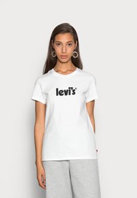 Camiseta de algodón blanca con el logo "levi's" en negro, de manga corta, con cuello redondo y corte recto; combinada con pantalones de chándal grises.