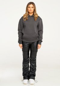 Grau Hoodie mit einer Fronttasche, kombiniert mit schwarzen Kunstlederhosen, die eng anliegen und sichtbare Nähdetails aufweisen. Weiße Sneaker runden den Look ab.