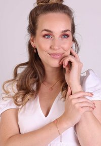 Sieradenset met goudkleurige schakels en hartvormige roze accenten. Model draagt een witte blouse en toont oorbellen en een halsketting.
