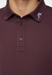 Burgunderröd polo skjorta i strukturerad tyg med klassisk krage, tre knappar och en liten vit logotyp på kragen.