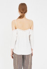White off-shoulder blouse with lace trim, three-quarter sleeves, and a gathered back. Model wears it with striped patterned trousers.