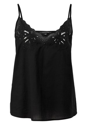Femme portant un haut noir sans manches avec des détails brodés et une jupe noire, debout, une main touchant un mur blanc.