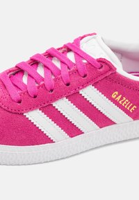 Vaaleanpunainen mokkanahkainen tennari, jossa valkoiset nahkaiset raidat, pyöreä kärki, valkoinen kumipohja ja keltaisella sivulla oleva "GAZELLE"-logo.