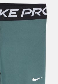 Grønne Nike Pro leggings med sort talje som har hvit "NIKE PRO" tekst og hvit Nike swoosh-logo på venstre lår.