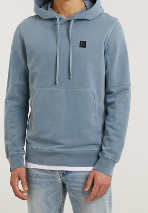 Mann trägt eine verblasste blaue Kapuzenjacke mit Fronttasche und Kordeln, kombiniert mit hellblauen Jeans vor einfarbigem Hintergrund.