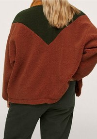 Fleecejakke i brændt orange og grøn med et chevron-design. Tekstureret stof, oversized pasform og brede ærmer. Afsluttet kant.