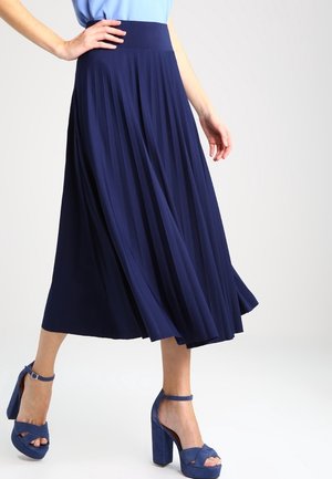 Kobieta w nosząca zwiewną granatową plisowaną spódnicę midi, połączoną z pasującymi niebieskimi sandałami na obcasie z paskiem wokół kostki, stojąca na jednolitym tle.