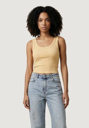 Donna con capelli ricci che indossa un top corto beige senza maniche e jeans a vita alta azzurri, in piedi contro uno sfondo semplice.