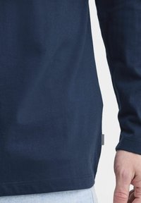 Granatowa koszula z długim rękawem o gładkiej bawełnianej teksturze, z boczną metką i zaokrąglonym dołem, pokazana z tyłu.