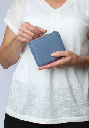 Portafoglio in pelle blu di forma quadrata, con chiusura a zip e dettagli di cucitura sottili. Tenuto in mano su uno sfondo chiaro.
