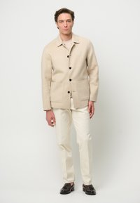 Giacca di lana beige con colletto, bottoni neri e due tasche frontali, abbinata a pantaloni crema chiara e mocassini scuri.