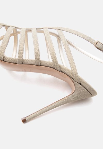 Μπεζ γυαλιστερό σανδάλι με λουράκια, μυτερή μύτη και λεπτό τακούνι στιλέτο σε λευκό φόντο.