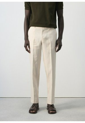 Modello maschile che indossa pantaloni beige su misura abbinati a sandali marrone scuro e una camicia a maniche corte verde scuro, in piedi su un pavimento grigio.
