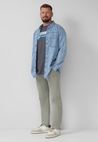 Man in een denim shirt, een donker graphic T-shirt, lichtgrijze broek en witte sneakers, staande met zijn handen in zijn zakken tegen een witte muur.