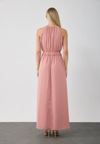 Maxiklänning i mjuk rosa tyg, halterneck-design, rynkad midjedetalj, flytande silhuett som faller till anklarna, bärs med beige klackar.