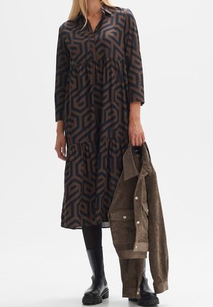 Donna che indossa un vestito midi con motivo geometrico, leggings neri, stivali neri robusti e tiene una giacca in camoscio marrone nella mano destra.