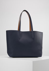 Sac fourre-tout bleu marine avec deux longues poignées, surface texturée et doublure intérieure marron sur les poignées, exposé sur un piédestal blanc.