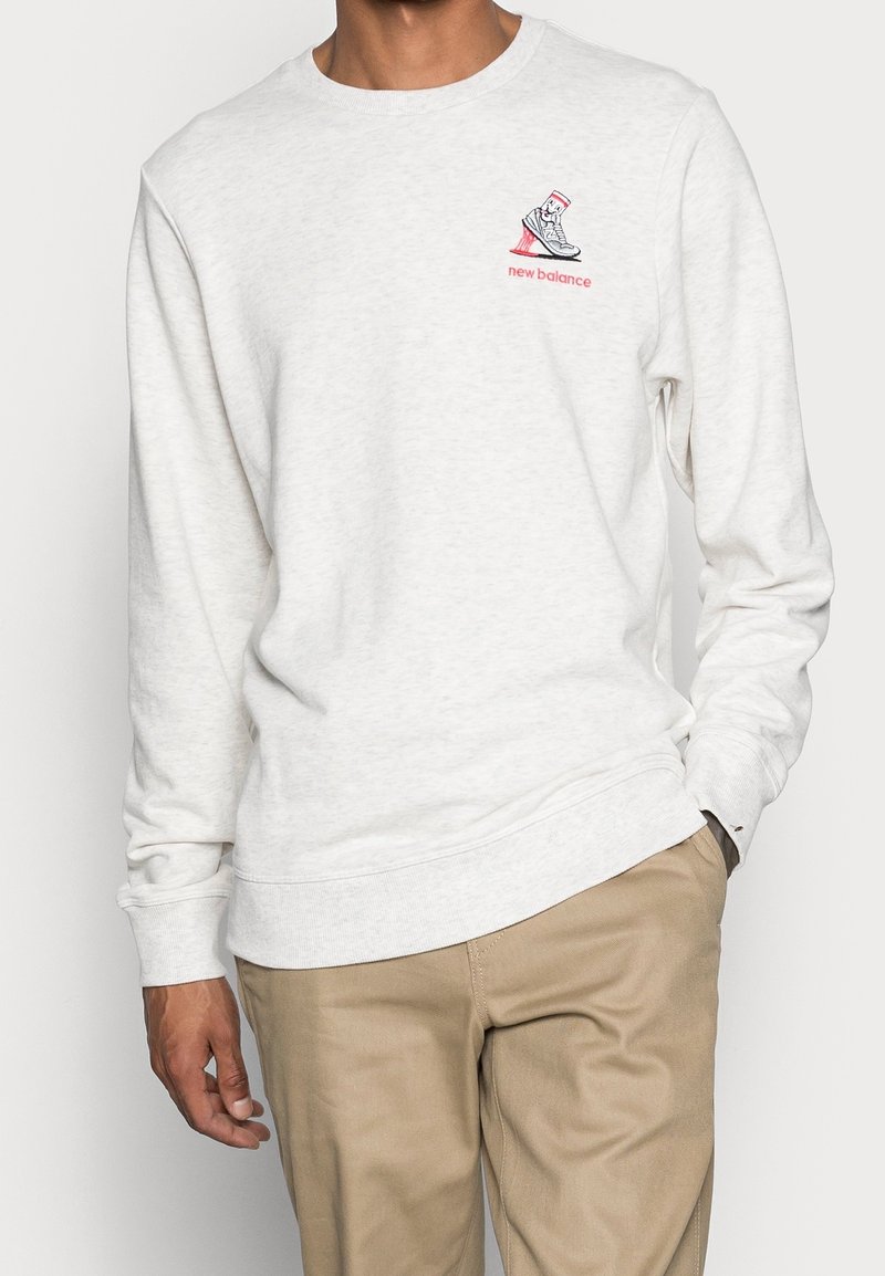 Sweat-shirt gris clair en tissu doux ; présente un logo avec un design de basket en rouge et noir sur le côté gauche de la poitrine. Manches longues et col rond.