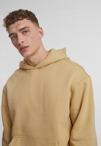 Felpa beige con cappuccio, realizzata in un morbido tessuto testurizzato e dotata di una tasca frontale. Il design presenta una vestibilità rilassata e spalle scese.