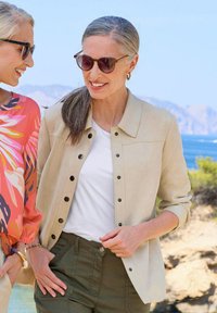 Veste en daim beige à boutons portée sur un t-shirt blanc, associée à un pantalon vert olive. Deux femmes, l’une portant un haut floral, à l'extérieur près d'une côte.