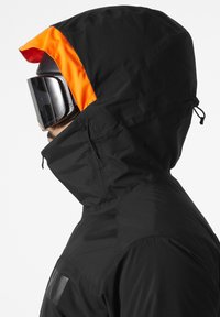 Veste de ski noire avec une doublure de capuche orange, poche latérale zippée et texture lisse. Comprend une capuche ajustable et un design épuré.