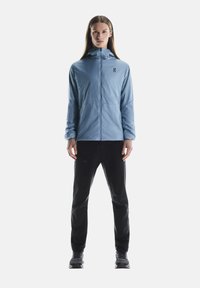 Veste zippée bleu clair avec capuche, arborant un logo sur la poitrine, associée à un pantalon noir et des chaussures foncées. Le tissu semble lisse et léger.