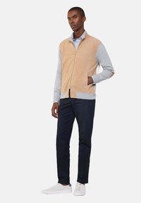 Beige Wildleder-Zip-Jacke mit grauen Ärmeln, Fronttaschen und einem Kragen. Getragen über einem hellblauen Hemd mit Knöpfen und einer navyfarbenen Hose.