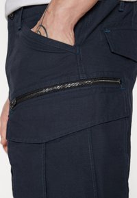 Pantaloni cargo navy din țesătură texturată, cu un buzunar lateral cu fermoar negru și detalii de cusătură albastră contrastantă.