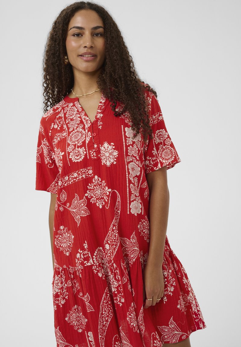 Frau mit langen lockigen Haaren, die ein rotes kurzes Kleid mit weißen Blumen- und Paisleymustern trägt, steht vor einem schlichten Hintergrund.