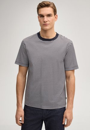 Junger Mann, der ein schwarz-weiß gestreiftes T-Shirt mit kurzen Ärmeln und dunkle Hosen trägt, steht vor einem schlichten hellen Hintergrund.