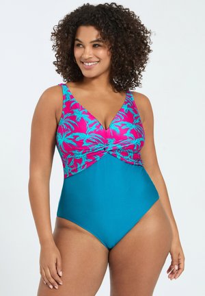 Donna sorridente con capelli ricci che indossa un costume intero con la parte superiore con motivo di palme rosa e blu e la parte inferiore blu unita.