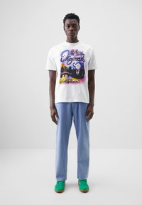 Weißes Baumwoll-T-Shirt mit buntem Grafikdesign, das Text und eine Figur zeigt. Kombiniert mit hellblauen gemusterten Hosen und grünen Sneakern.