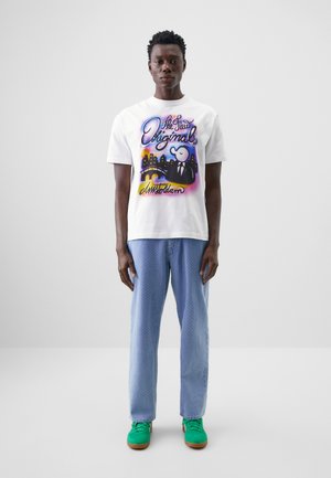 White cotton t-shirt featuring colorful graphic design with text and a character. Paired with light blue patterned trousers and green sneakers.
