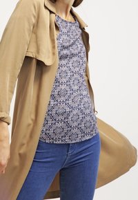 Leggera giacca beige su una maglietta blu a fantasia con motivi floreali. Jeans blu aderenti visibili, creando un look a strati.