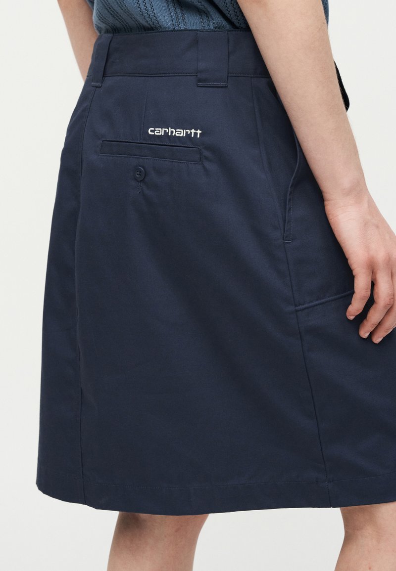 Gonna blu navy al ginocchio con passanti per cintura, tasca posteriore con bottone, tasca laterale e logo Carhartt bianco sopra la tasca posteriore.
