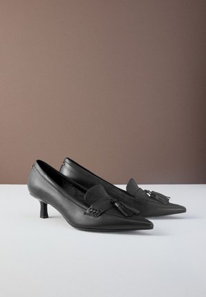 Schwarze High-Heel-Schuhe aus Leder mit spitzer Zehenpartie und Quastendetail vorne. Glatte Textur und ein geometrisch gestalteter Absatz.