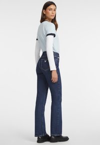 Camiseta azul claro con detalles en azul marino, superpuesta a una camisa blanca de manga larga. Pantalones de mezclilla oscuros de pierna acampanada con un logo triangular en la parte posterior.