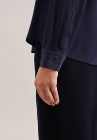 Marineblauwe overhemd met lange mouwen en een knoopmanchet, met een gladde textuur en een ontspannen pasvorm, gedeeltelijk weergegeven met een hand die aan de zijkant rust.