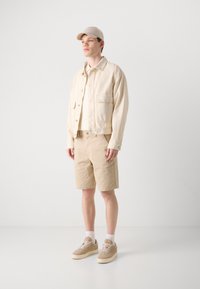 Beige džínsová bunda, krémová košile, khaki šortky a béžové tenisky. Jednoduchý design, zapínací bunda s kapsou na náprsní části, nošena s baseballovou čepicí.