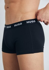 Schwarze Boxershorts mit enger Passform, ausgestattet mit einem breiten Bund, auf dem "HUGO" in Weiß gedruckt ist. Das Material wirkt glatt und dehnbar.