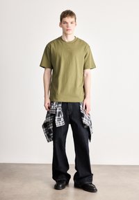 Bombažna majica barve oljčne zelene z okroglim izrezom, kombinirana s temno modrimi širokimi kavbojkami in črno-belo karo flanelasto srajco, privezano okoli pasu.