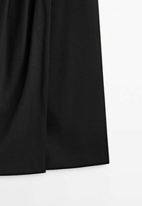 Détail d'une jupe en tissu noir montrant une texture lisse et de doux plis sur un fond blanc uni.
