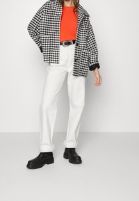 Veste oversize à motif pied-de-poule noir et blanc portée sur un top orange vif, associée à un jean blanc à jambes larges et des bottines noires épaisses.
